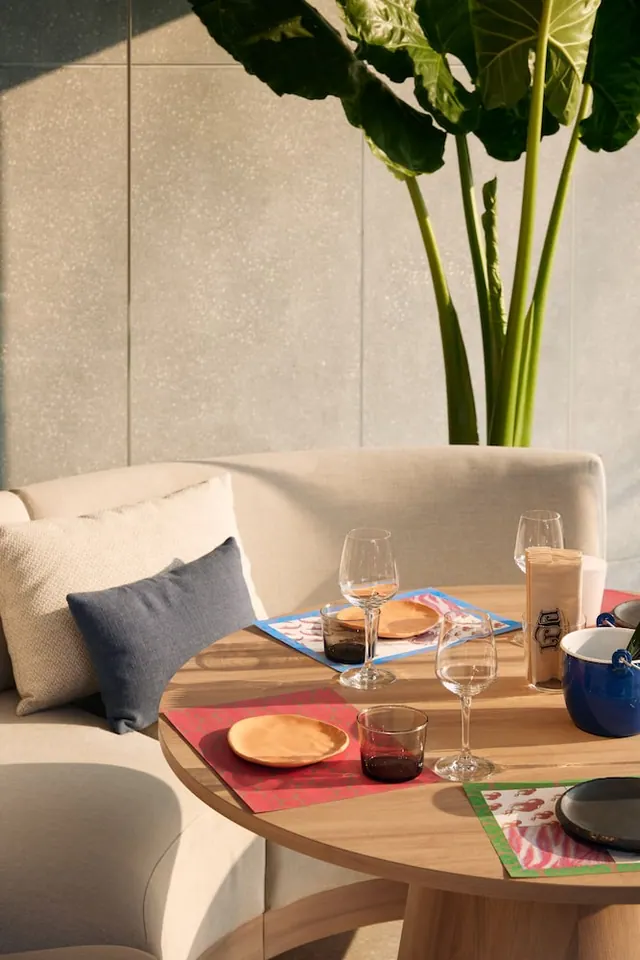 A round wooden table is set with wine glasses, empty plates and placemats beside a cushioned sofa and a large potted plant against a light stone wall.