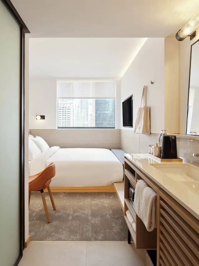 A hotel room with a made white bed beside a window showing high-rise buildings, a wall-mounted television, and a wooden vanity with a sink, kettle and hanging towels.