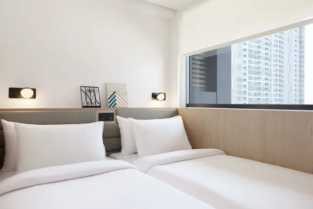 Two beds with white bedding and pillows sit beside a window that looks out onto high-rise buildings.