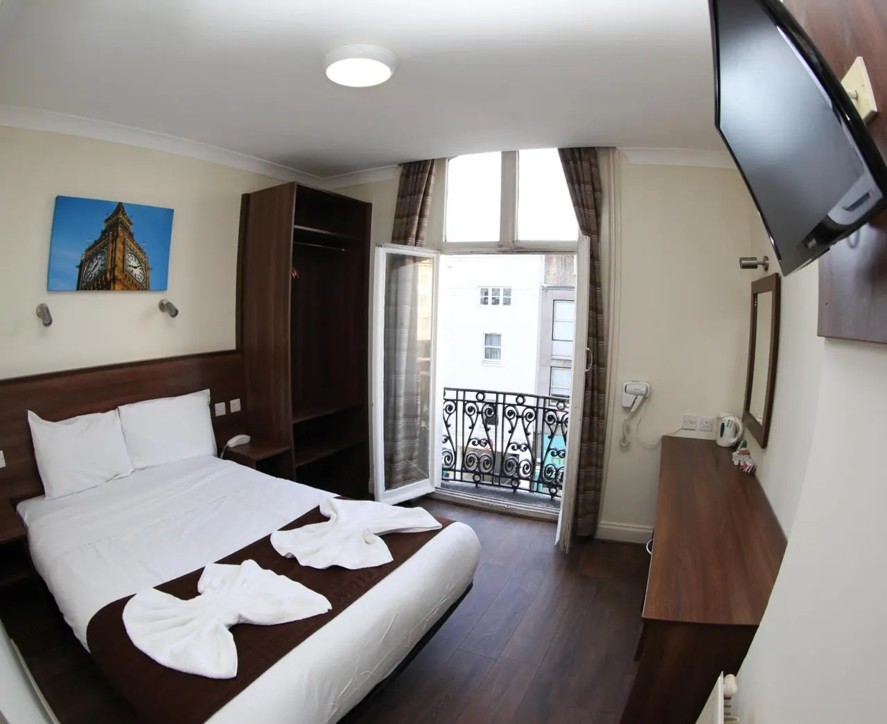 A double bed with white towels folded into animal shapes sits beside a wardrobe, wall-mounted television and a balcony door that opens onto a view of a building opposite.