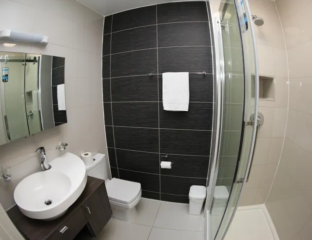A bathroom with a white vessel sink on a dark wood vanity, a toilet, a black tiled feature wall with a hanging towel, and a glass-enclosed shower.
