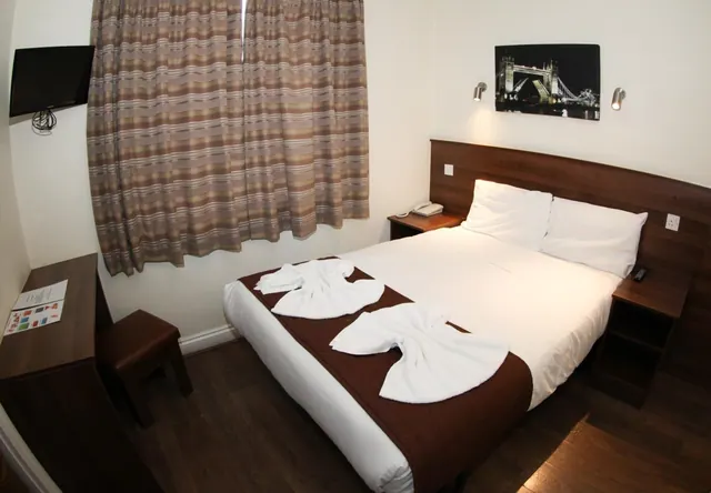 A double bed with white linen and two folded towels sits beside a window with striped curtains, a wall-mounted television, and a framed print of Tower Bridge.