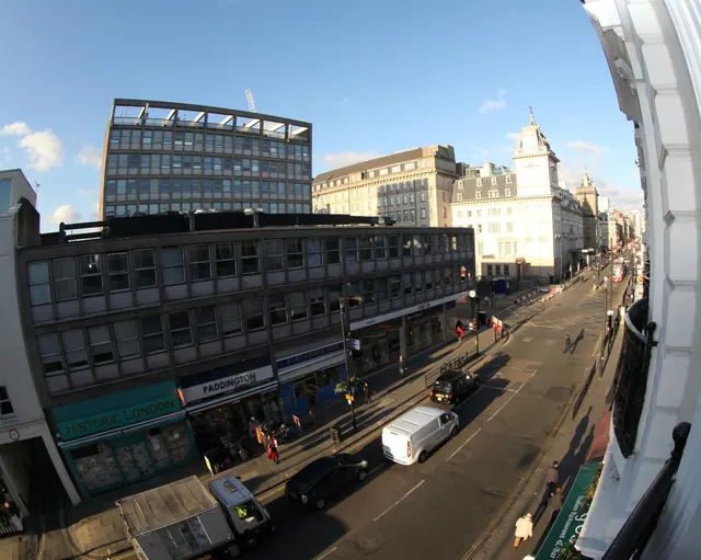 Photo taken from an upper floor balcony looking down a wide city street lined with multi-storey buildings, shop fronts, and parked and moving vehicles.