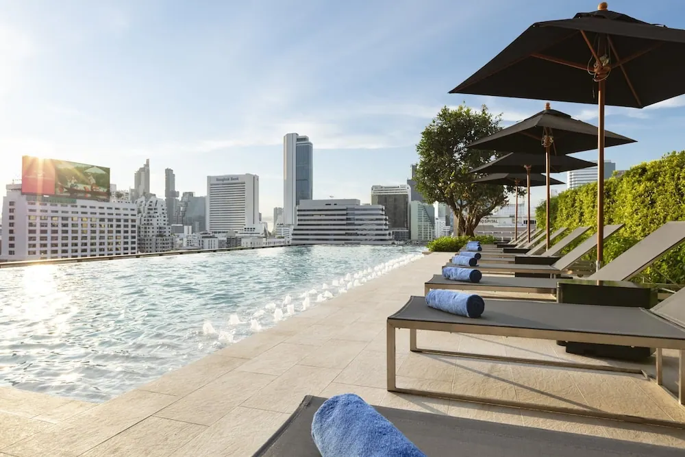 A rooftop infinity pool with sun loungers, shade umbrellas and a view of high-rise buildings under a clear sky.