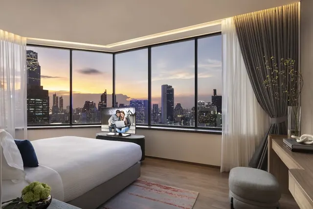 A bedroom with a large bed, a television on a low cabinet, a wooden desk, and floor-to-ceiling windows showing a city skyline at dusk.