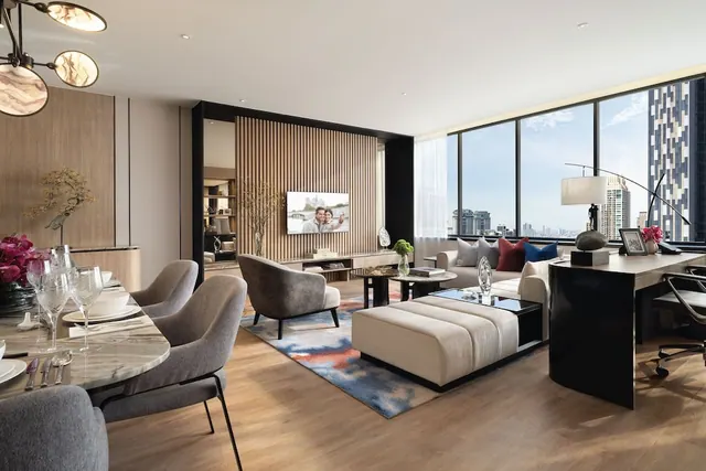 A staged living room with a sofa, ottoman, armchair, wall-mounted television, dining table and desk, with floor-to-ceiling windows overlooking high-rise towers.