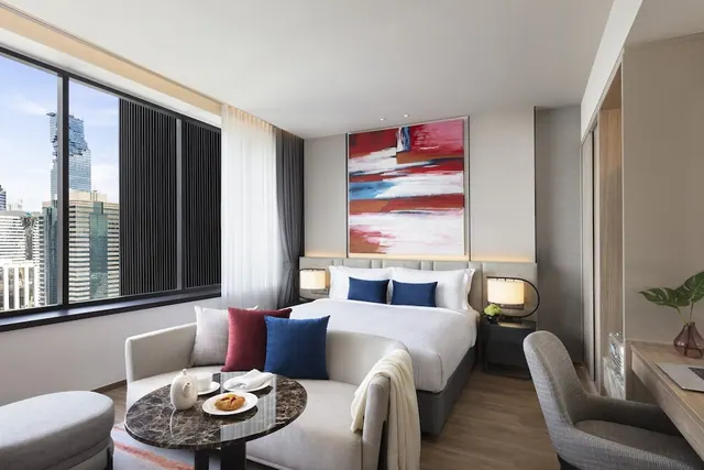A hotel bedroom with a king bed, a sofa with a round marble table, and a large window showing a city skyline.