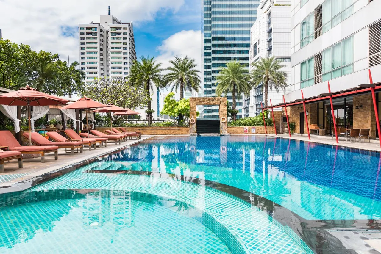 An outdoor swimming pool with blue tiles, orange sun loungers and pink umbrellas, with tall towers behind the hotel building.