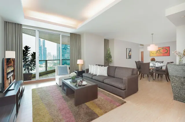 A living room with a grey sofa, armchair, coffee table and wall-mounted television, with sliding doors onto a balcony showing city towers outside.