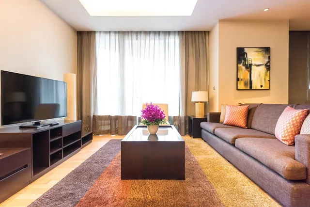 A living room with a flat-screen television on a dark media console, a grey sofa with patterned cushions, and a coffee table with a purple flower arrangement on a striped rug.