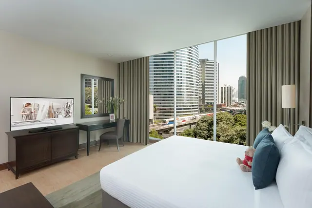 A bedroom with a made king bed, a television on a dark wooden cabinet, a desk with a chair, and large windows looking onto high-rise buildings and an elevated train line.