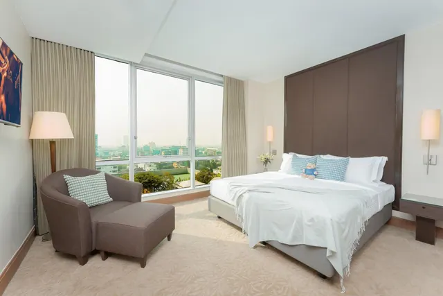A bedroom with a king bed, an armchair with an ottoman, and floor-to-ceiling windows overlooking a city with trees and low buildings.