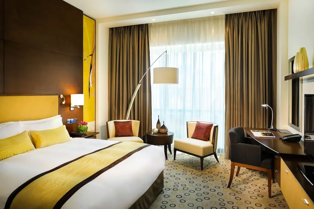 A hotel bedroom with a king bed, two armchairs, a floor lamp, a desk and a television beside sheer-curtained windows.
