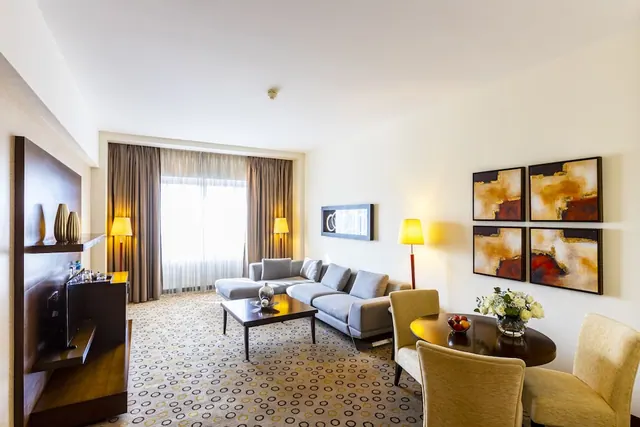 A living area in a hotel suite with a grey sectional sofa, a coffee table, floor lamps, a round dining table with flowers and fruit, and four abstract paintings on the wall.
