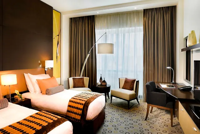 A hotel room with two single beds with orange and brown throws, two armchairs, an arc floor lamp, and a desk with a telephone beside a window with sheer curtains.