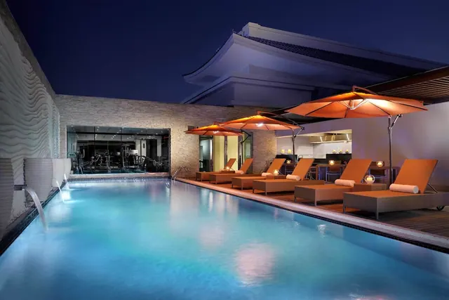 An illuminated outdoor pool at night with orange parasol umbrellas and sun loungers along the edge, and a gym visible through a glass wall behind.