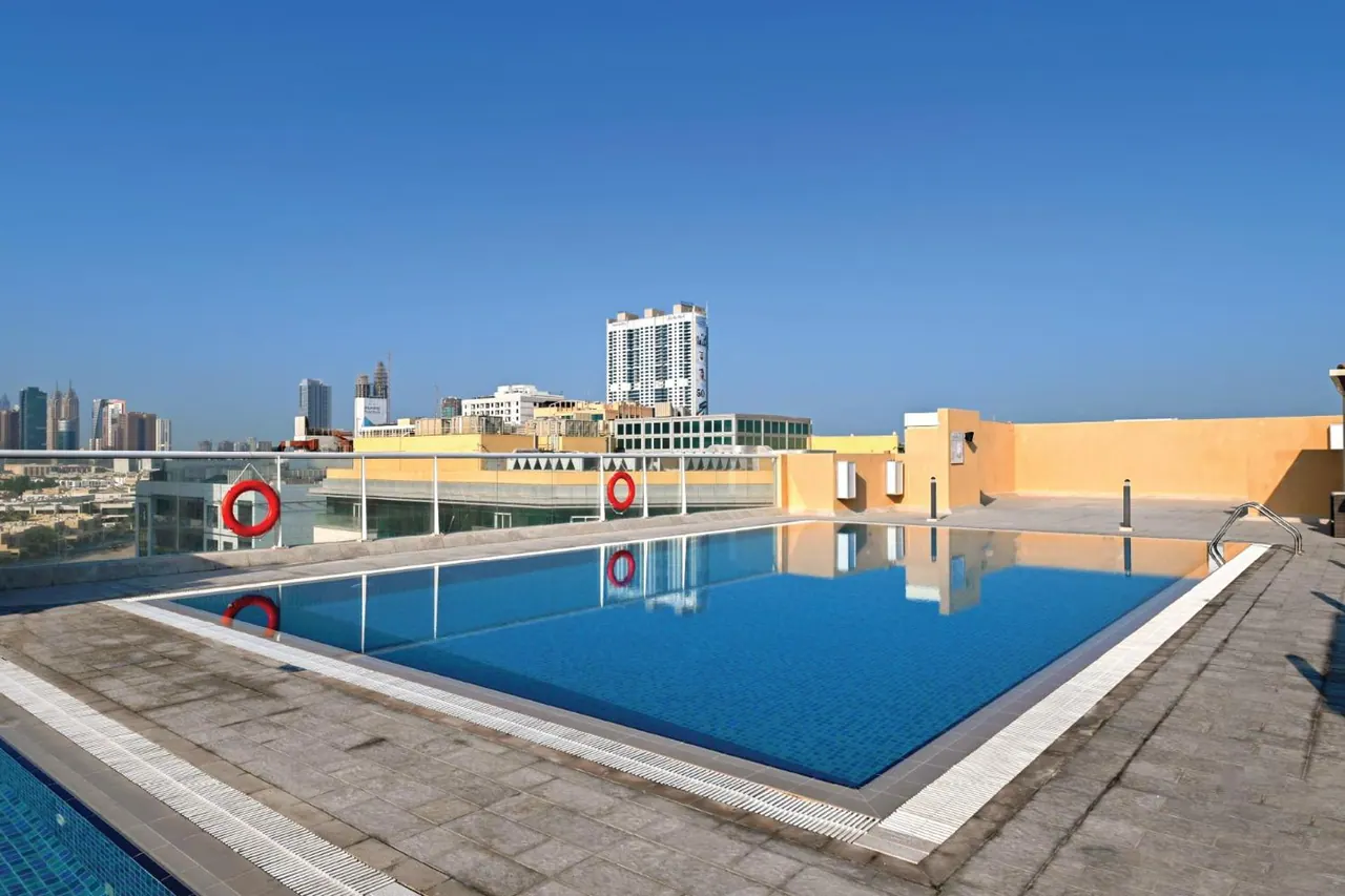 A rooftop swimming pool with red life rings on the railing, with a city skyline and a clear blue sky behind it.