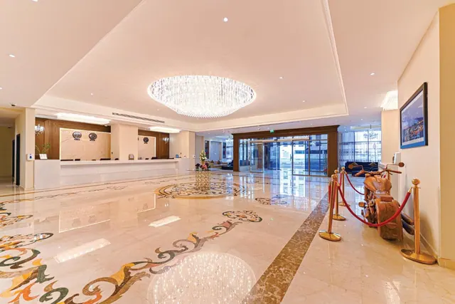 A large marble-floored hotel lobby with a crystal chandelier, a reception desk on the left, glass entrance doors ahead and a wooden motorcycle sculpture behind red rope barriers on the right.