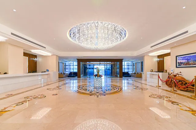 The hotel lobby has a large crystal chandelier, a marble floor with medallion inlays, reception desks on both sides, and sofas near the glass entrance doors.