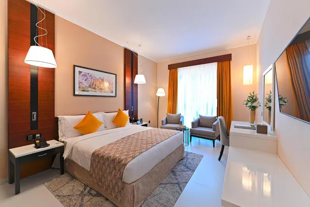 A hotel bedroom with a king bed, two armchairs beside a window with orange curtains, and a wall-mounted television.