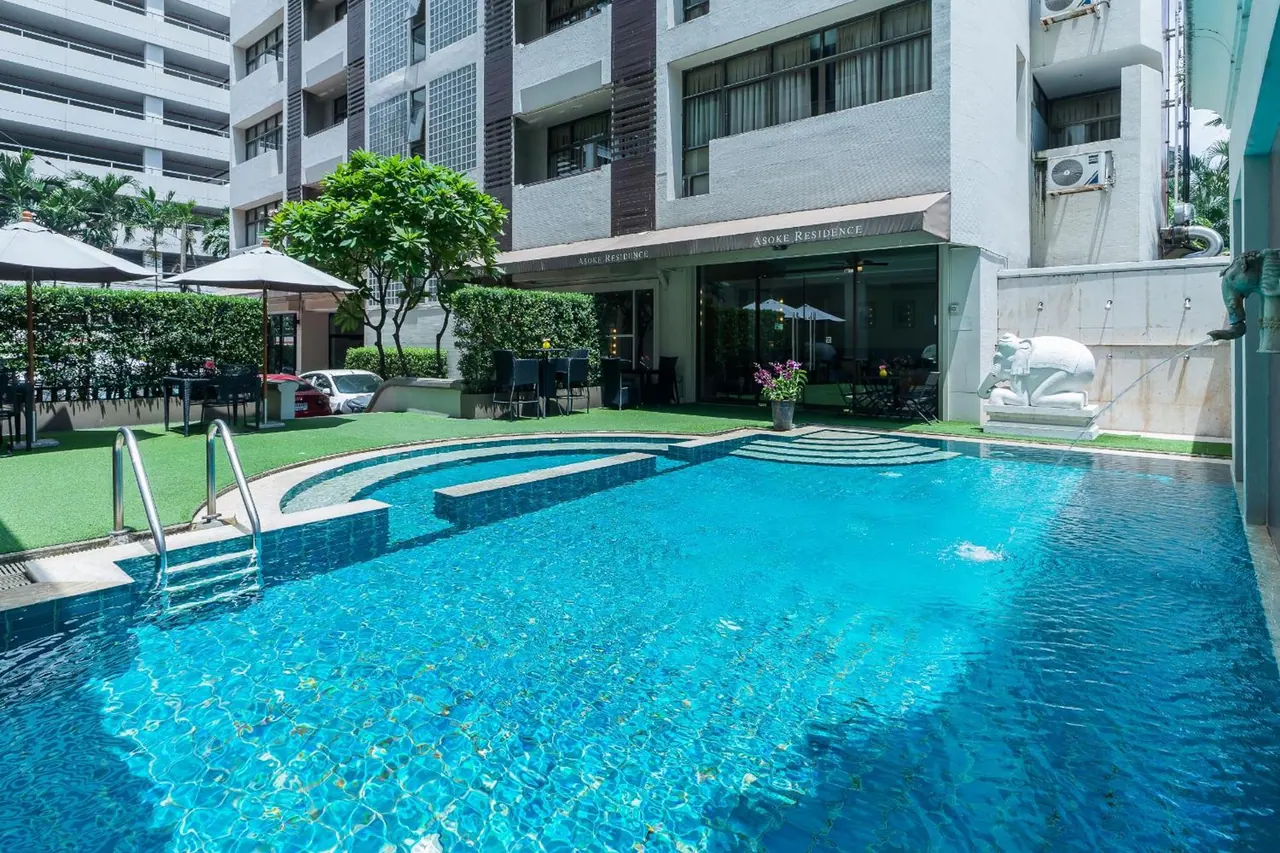 A blue-tiled swimming pool with entry steps and a ladder sits in front of a white multi-storey building, with outdoor tables, chairs and parasols on the lawn beside it.