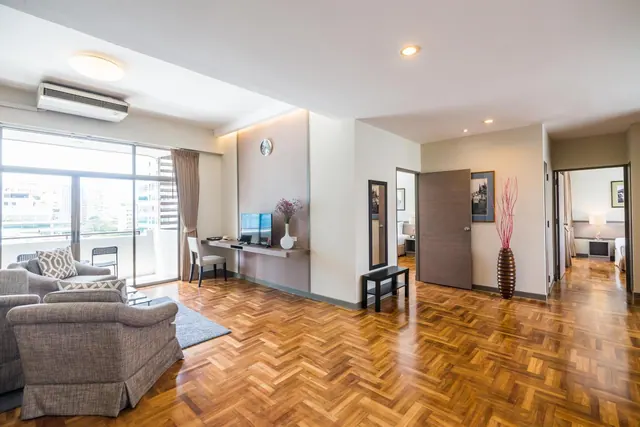A suite living area with grey armchairs, a wall-mounted television and desk, parquet flooring, and sliding glass doors onto a balcony with city buildings beyond.