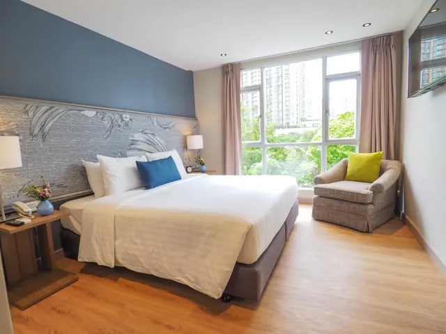 A bedroom with a made king bed, a blue headboard wall, an armchair with a green cushion, and a large window overlooking high-rise buildings and trees.