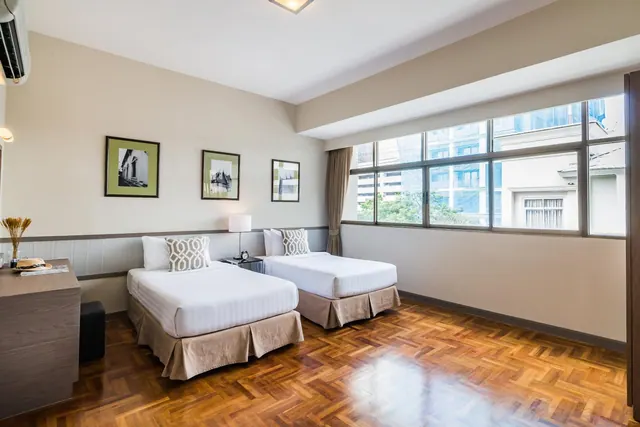 A bedroom with two twin beds with white bedding, framed prints on the wall, a wooden dresser on the left and a wide window looking out onto neighbouring buildings and trees.