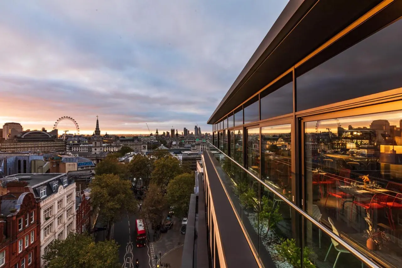A glass-walled terrace overlooks central London at sunset, with the London Eye, a church spire and a red bus on the street below.