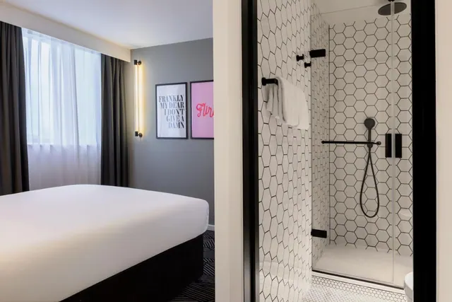 A made bed sits beside a curtained window and framed wall prints, while a glass-doored walk-in shower with hexagon-tiled walls is visible to the right.