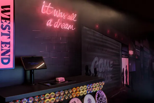 A dark, themed reception area with a pink neon sign reading 'it was all a dream', a check-in screen on a counter studded with skateboard wheels, and a painted wall reading 'Come Alive'.
