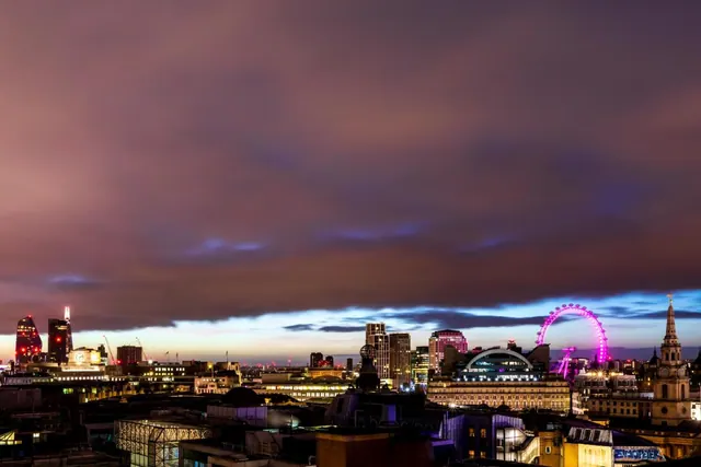 Dusk view over a London skyline with the illuminated London Eye, a church spire and office towers under a cloudy sky.