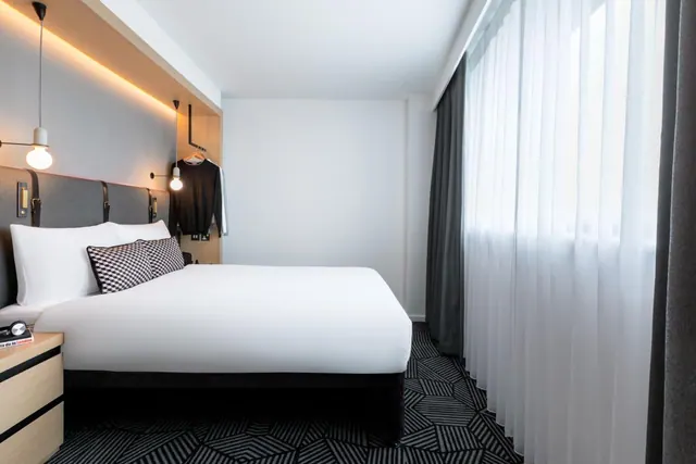 A bedroom with a double bed under white bedding and patterned cushions, wall lights above the headboard, a hanging clothes rail, and sheer curtains at a window.