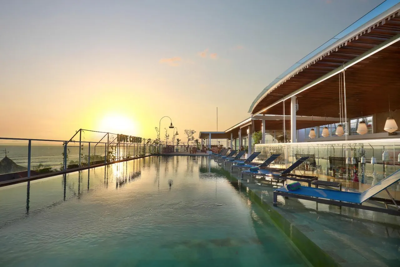 An infinity pool with sun loungers beside a covered terrace at sunset, with the sea visible beyond the railing.