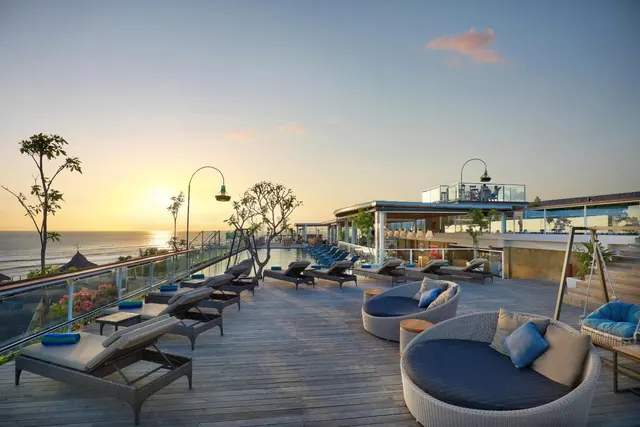 A wooden poolside terrace at sunset with sun loungers, round outdoor daybeds and a view over the ocean.
