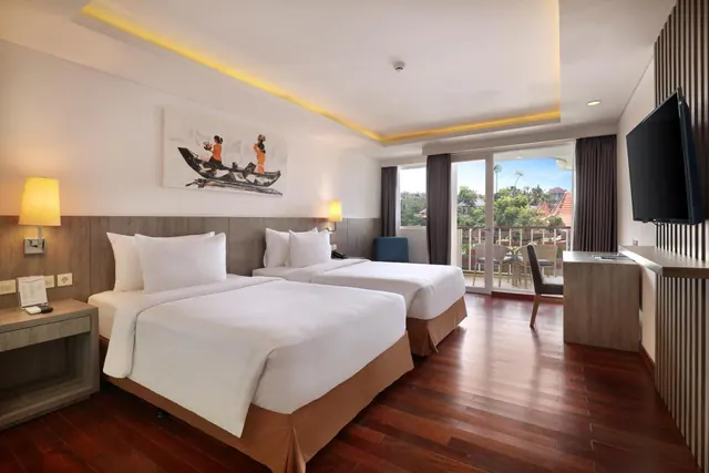 A twin-bedded hotel room with a wall painting, a wall-mounted television, a desk, and a balcony with sliding glass doors looking onto trees and rooftops.