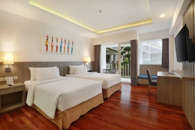 A bedroom with two beds, a wall-mounted television, a desk and chair, and an open glass door onto a balcony with plants outside.