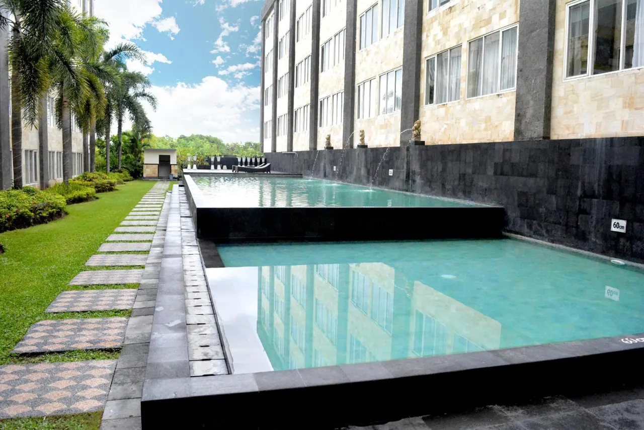 A long pool with wall-fed water features runs beside a hotel building, with palm trees and a grass path along one side.
