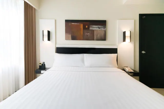 A bedroom with a made-up king bed, white pillows, a black headboard, wall sconces, an abstract painting and a black door.
