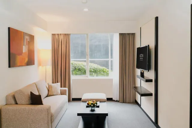 A living area with a beige sofa, a low black coffee table with a flower arrangement, a wall-mounted television and a window looking out onto green shrubs.