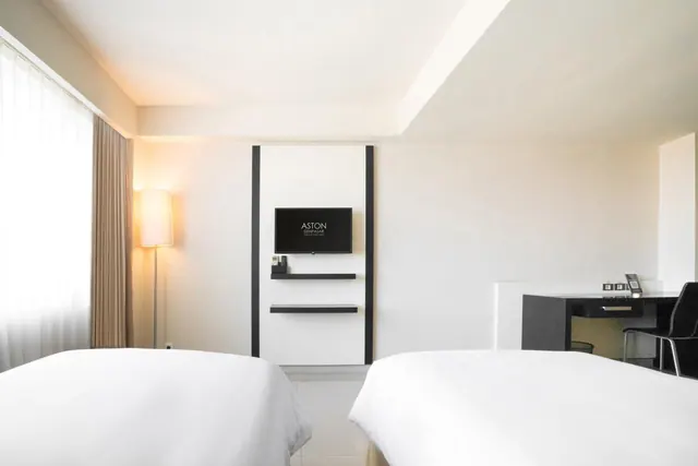 A hotel bedroom with two white beds, a wall-mounted television, a floor lamp by the window and a desk with a chair.