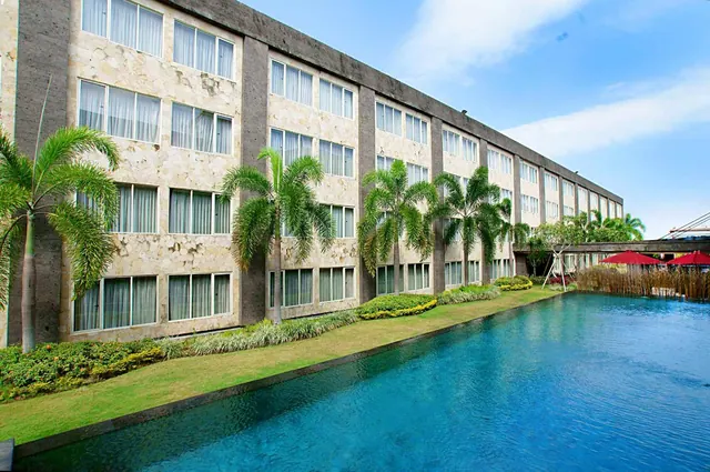 A long multi-storey hotel building with a stone-clad facade and palm trees stands beside a long swimming pool, with red umbrellas visible on the right.