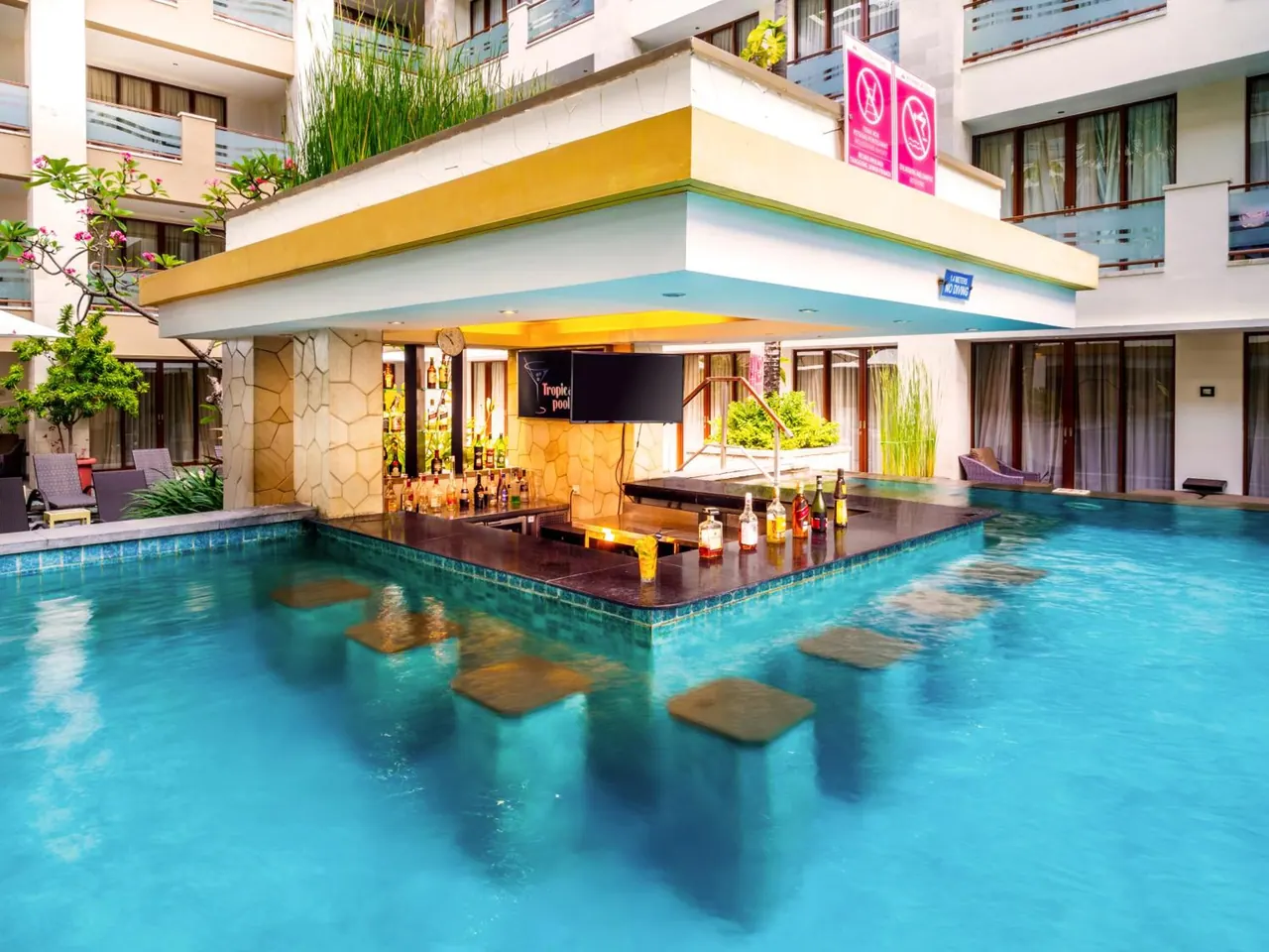 A swim-up bar with bottles and a wall-mounted TV stands in a turquoise pool, with hotel building walls and balconies behind it.