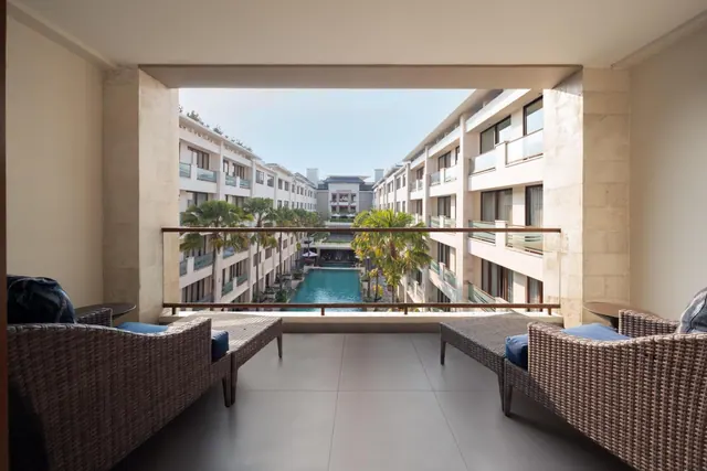 A covered balcony with two wicker armchairs and blue cushions looks out over a courtyard pool surrounded by hotel buildings and palm trees.