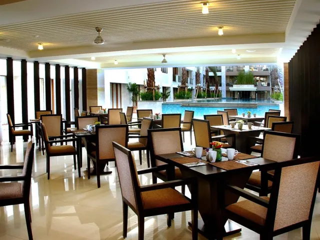 A dining room with set tables and upholstered chairs, with a swimming pool, palm trees and hotel buildings visible through the open side.