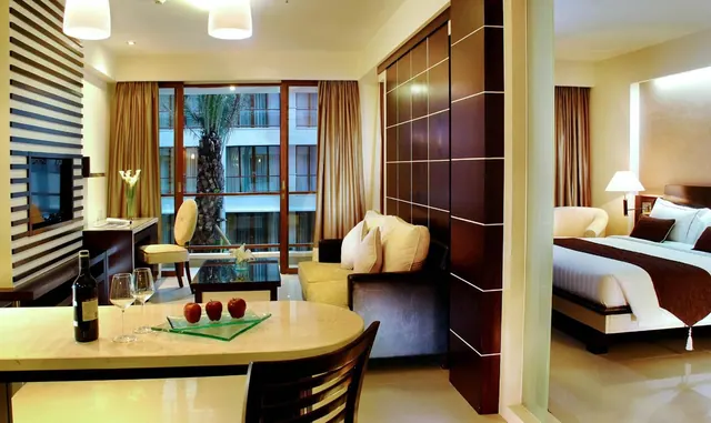 A hotel suite living area with a dining table holding a wine bottle, two glasses and apples, a sofa and glass coffee table, a view through glass doors of a palm tree and a building, and a bed visible in the adjoining bedroom.