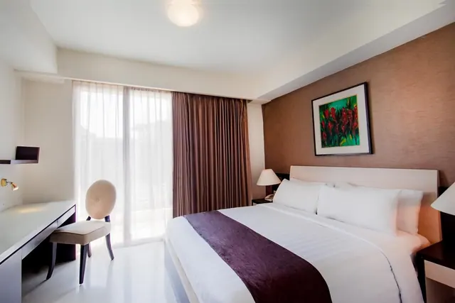 A bedroom with a made king bed, a desk and chair beside sheer-curtained glass doors, and a framed painting above the headboard.