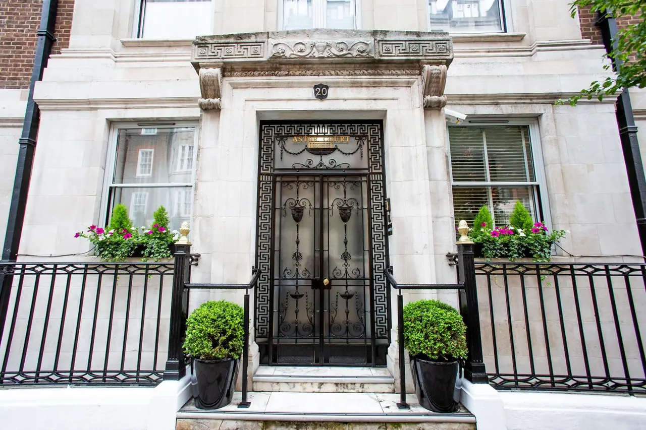 The stone facade of the building has an ornate iron entrance gate, a number 20 plaque, and planters with topiary and flowers beside the steps.