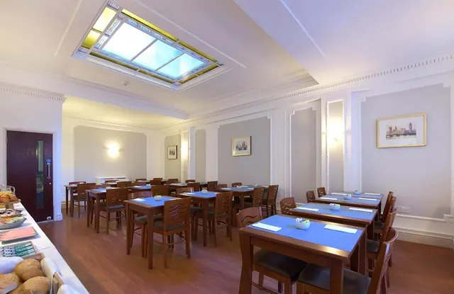 A dining room with blue-clothed tables and wooden chairs under a stained glass skylight, with a breakfast buffet counter with bread along the left edge.