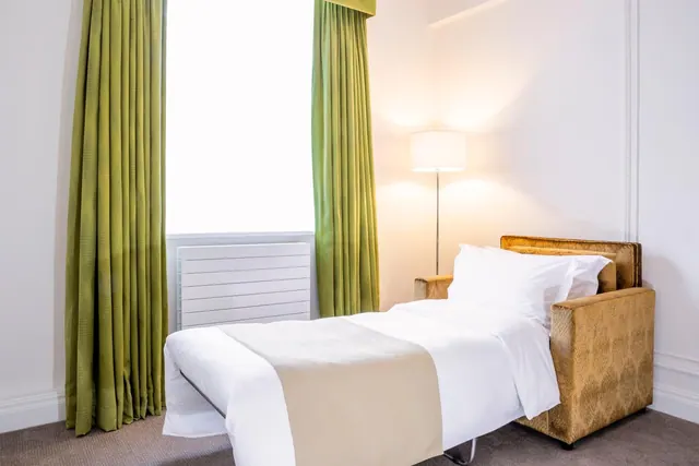 A made-up sofa bed with white bedding and a beige throw sits beside green curtains, a floor lamp and a bright window in a bedroom.